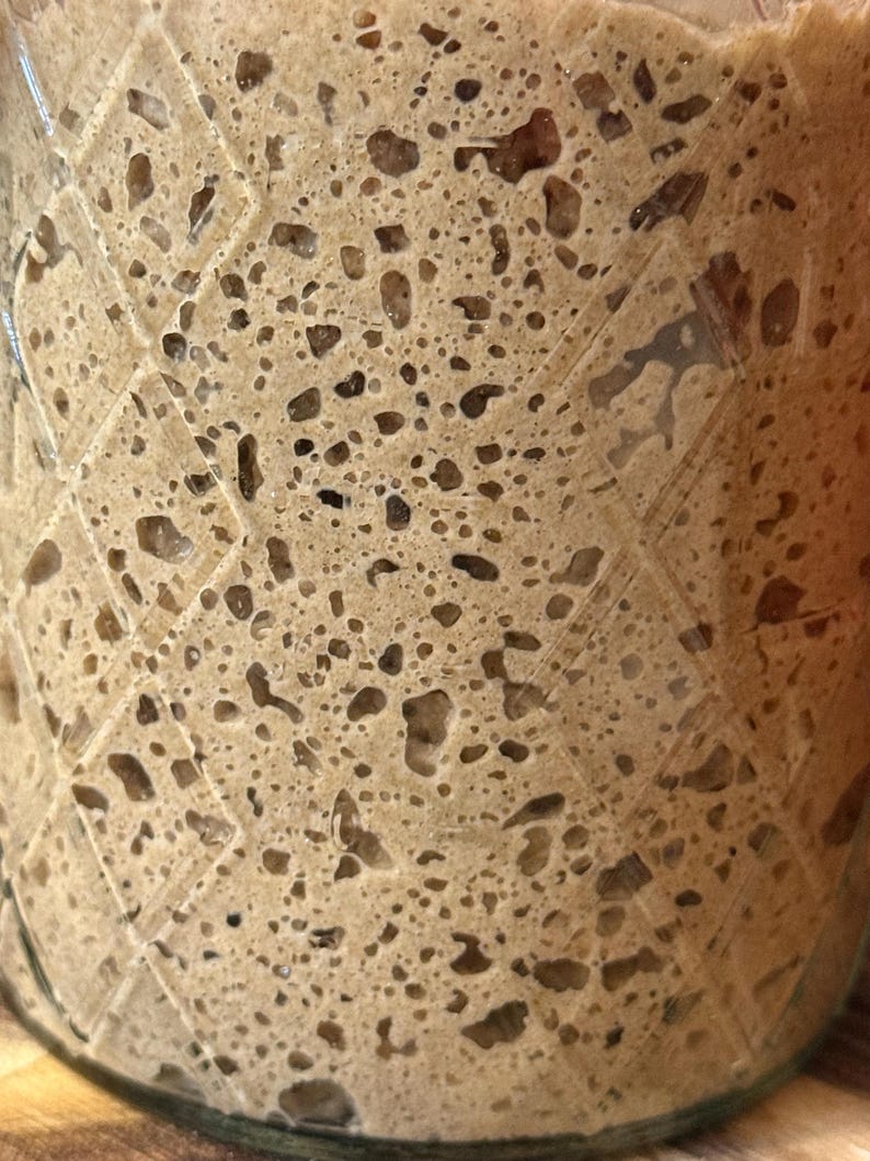 May include: Close-up of a glass jar filled with a light brown, bubbly mixture. The mixture has a porous texture with numerous air pockets and darker brown inclusions. The jar has a diamond pattern etched into the glass.