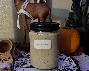 May include: A glass jar with a black lid filled with a light brown sourdough starter labeled "New Zealand Rye". The jar is sitting on a black and white patterned plate.