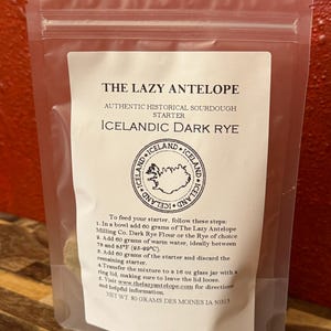 May include: A transparent packet of sourdough starter, labelled "THE LAZY ANTELOPE" and "ICELANDIC DARK RYE." The label includes instructions for use and a logo. The net weight is 80 grams. The instructions include measurements in grams and temperature in Celsius.