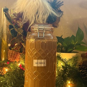 Könnte beinhalten: Ein dekoratives Glas mit einem Klappdeckel, gefüllt mit einer braunen Substanz. Das Glas hat ein strukturiertes Rautenmuster und ein Etikett mit der Aufschrift "RUSSIA". Das Glas ist von Weihnachtsdekorationen umgeben.