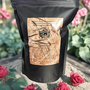 May include: A black bag of fresh milled stone-ground Einkorn flour. The label reads "Fresh Milled Stone-Ground Einkorn Flour" and "The Mother of All Wheat". The bag is surrounded by red roses and a blurred background of pink flowers.