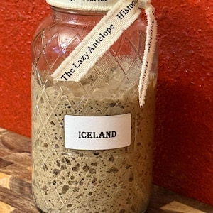 Könnte beinhalten: Ein Glas mit Sauerteigstarter, beschriftet mit "Iceland". Das Glas ist mit einem Band mit der Aufschrift "Sourdough Starter" und "The Lazy Antelope Histor" verziert. Das Glas steht auf einer Holzoberfläche vor rotem Hintergrund.