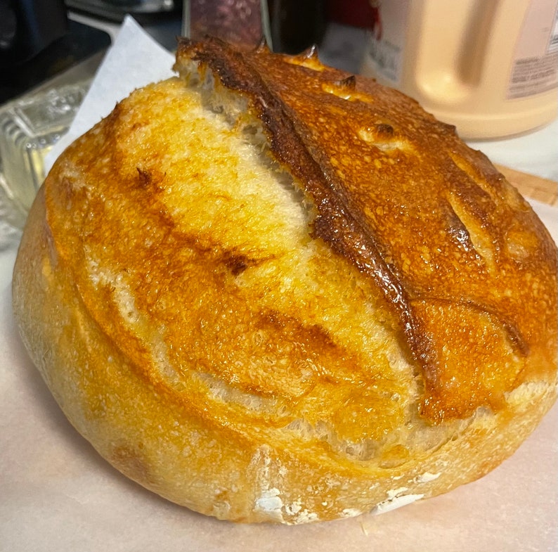 Op de afbeelding: Een vers gebakken zuurdesembrood met een goudbruine korst. Het brood heeft een licht onregelmatige vorm en een zachte, luchtige textuur.