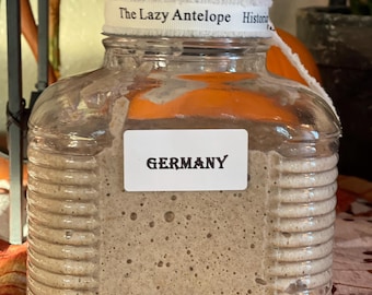 May include: A clear glass jar with a white label that reads "GERMANY". The jar is filled with a light brown, bubbly substance. The jar has a white ribbon around the top that reads "The Lazy Antelope Historic".