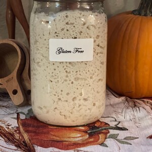 May include: A glass jar filled with a white, bubbly substance labeled "Gluten Free". The jar is sitting on a table with a white tablecloth with a pumpkin design.