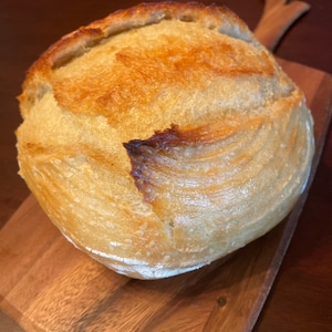 May include: A freshly baked loaf of sourdough bread with a golden brown crust and a slightly cracked surface. The bread is sitting on a wooden cutting board.