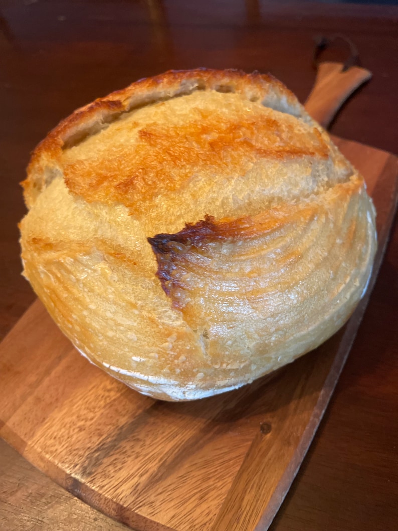 May include: A freshly baked loaf of sourdough bread with a golden brown crust and a slightly cracked surface. The bread is sitting on a wooden cutting board.