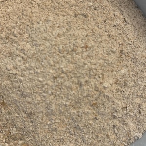 May include: Close-up of a pile of light brown, grainy flour. The texture appears coarse and uneven, with visible flecks and particles. The flour is likely for baking or cooking.