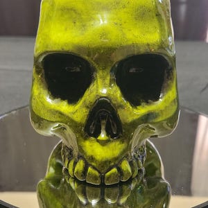 May include: A bright green ceramic skull mug with black eye sockets and teeth. The mug is sitting on a reflective surface.
