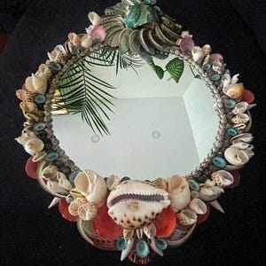 May include: A round mirror with a decorative frame made of various seashells. The frame is a mix of colors, including white, brown, pink, and blue. The mirror is surrounded by a variety of seashells, including cowrie shells, clam shells, and scallop shells.