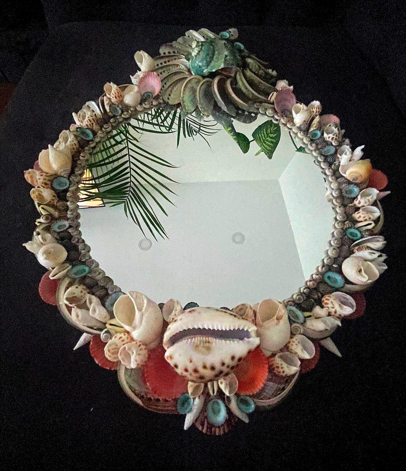 May include: A round mirror with a decorative frame made of various seashells. The frame is a mix of colors, including white, brown, pink, and blue. The mirror is surrounded by a variety of seashells, including cowrie shells, clam shells, and scallop shells.