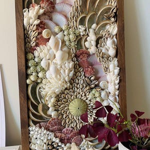 Peut inclure: Une œuvre d'art en mosaïque de coquillages encadrée avec une variété de coquillages dans des tons de blanc, de brun et de rose. Les coquillages sont disposés en un motif tourbillonnant, créant un design unique et accrocheur.