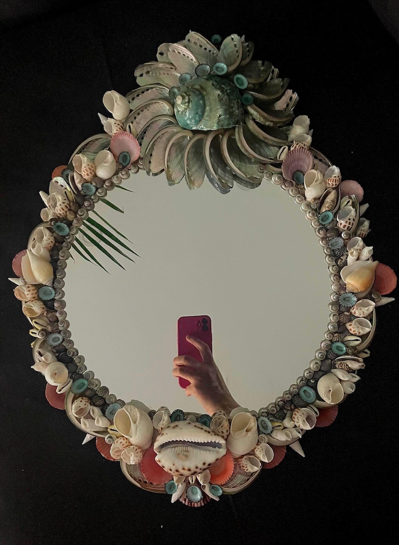 May include: A round mirror with a decorative frame made of various seashells. The frame is a mix of white, pink, and brown shells, with some blue and green accents. The mirror is reflecting a hand holding a pink phone.