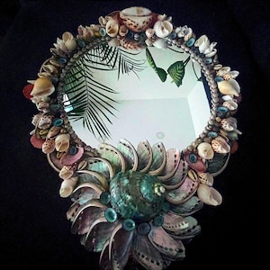 May include: A round mirror decorated with a variety of seashells in shades of pink, white, brown, and green. The mirror is surrounded by a border of shells, and the center features a large, iridescent shell.