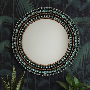 May include: Circular decorative mirror with a beaded frame. The frame has a pattern of teal and gold-coloured beads. The mirror is mounted on a dark background with a tropical leaf pattern. Home decor.