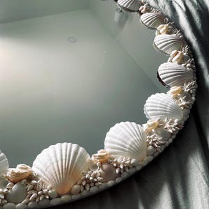 May include: Circular mirror with a decorative border of seashells. The shells are mainly white and cream, with smaller shells and pebbles filling the spaces. The mirror reflects the surrounding area, creating a coastal theme.