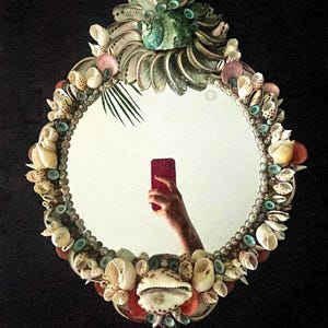 May include: A round mirror with a decorative frame made of seashells. The frame is made of a variety of shells in different colors and sizes, including white, brown, pink, and green. The mirror is oval-shaped and has a silver backing.