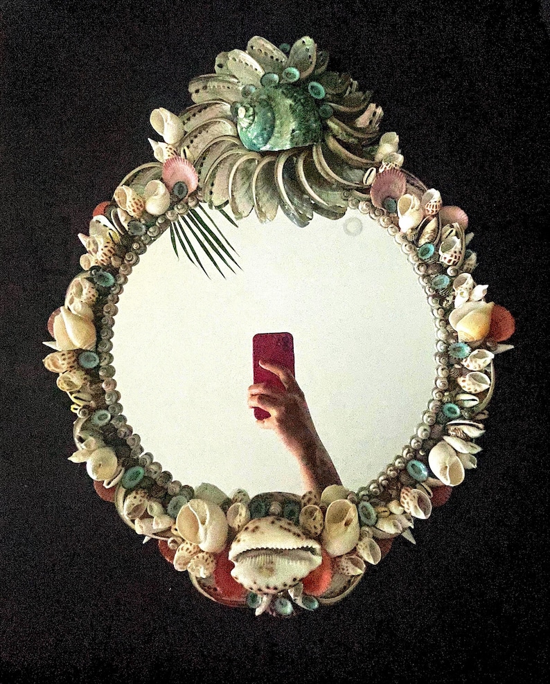May include: A round mirror with a decorative frame made of seashells. The frame is made of a variety of shells in different colors and sizes, including white, brown, pink, and green. The mirror is oval-shaped and has a silver backing.