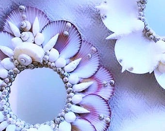 May include: Two round mirrors decorated with seashells. The mirror on the left is surrounded by white and purple seashells. The mirror on the right is surrounded by white seashells.