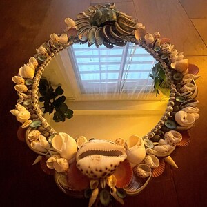 May include: A round mirror with a decorative frame made of seashells. The frame is made of a variety of shells in different colors and sizes, including white, brown, and pink. The mirror is reflecting a window with blinds.