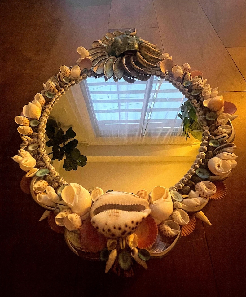 May include: A round mirror with a decorative frame made of seashells. The frame is made of a variety of shells in different colors and sizes, including white, brown, and pink. The mirror is reflecting a window with blinds.