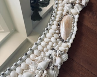 May include: A round mirror with a frame intricately decorated with various seashells, small white pebbles, and tiny silver beads. The seashells are primarily white and beige, creating a coastal or nautical theme.
