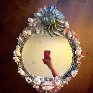 May include: A round mirror with a decorative frame made of seashells. The frame is a mix of white, pink, and blue shells. The mirror is reflecting a person taking a selfie with a pink phone.