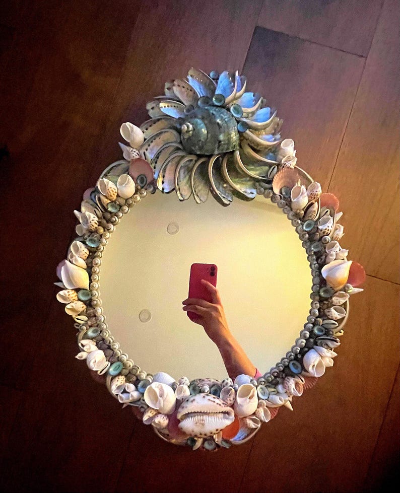 May include: A round mirror with a decorative frame made of seashells. The frame is a mix of white, pink, and blue shells. The mirror is reflecting a person taking a selfie with a pink phone.