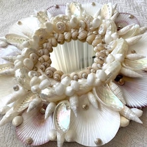 May include: A white seashell mirror with a round mirror in the centre. The mirror is surrounded by a variety of white seashells, including scallop shells, oyster shells, and small white shells.