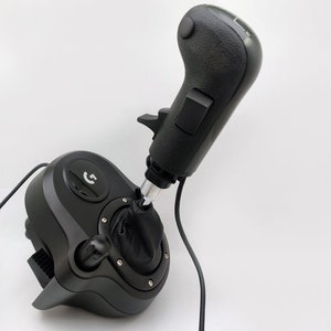 K&ouml;nnte beinhalten: Ein schwarzer Logitech G-Schalthebel mit einem Leder-Schaltknauf und einem silbernen Schaltstab. Der Schalthebel hat eine schwarze Basis mit einem Knopf und einem kleinen Hebel. Der Schalthebel ist mit einem schwarzen Kabel verbunden.