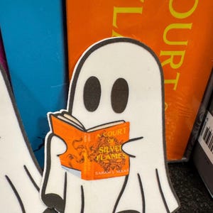 May include: A white ghost-shaped sticker reading a book with an orange cover. The book's title is "A Court of Silver Flames" by Sarah J. Maas. The ghost has black eyes and a black outline.