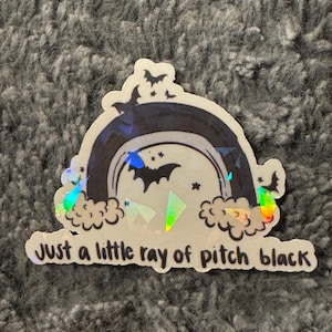 May include: A holographic sticker with a black and white rainbow, bats, and stars. The text "Just a little ray of pitch black" is printed on the sticker.