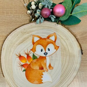 Könnte beinhalten: Ein Aquarellbild eines Fuchses auf einer Holzscheibe. Der Fuchs ist orange und weiß mit einem buschigen Schwanz. Der Hintergrund ist hellbraunes Holz.