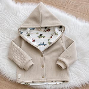 Könnte beinhalten: Eine beigefarbene Kapuzenjacke aus Waffelstrick mit langen Ärmeln und drei Knöpfen auf der Vorderseite. Das Innenfutter besteht aus weißem Stoff mit einem Muster aus kleinen bunten Autos und Tieren. Ein kleines weißes Etikett mit Herz ist am Saum.