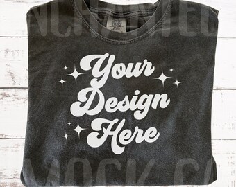 May include: A dark gray t-shirt with the text "Your Design Here" in white with small white stars on either side of the text. The shirt is folded and laid flat on a white wooden surface.