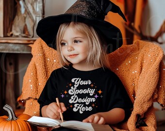 Può includere: Un bambino piccolo che indossa un cappello da strega nero e una maglietta nera con il testo "Your Spooky Design Here" in un font retrò. Il bambino è seduto su una coperta marrone e tiene un libro.