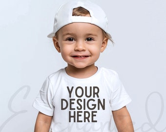Puede incluir: Una camiseta blanca para niños pequeños con mangas cortas y el texto "YOUR DESIGN HERE" impreso en marrón en la parte delantera. El niño lleva una gorra de béisbol blanca.