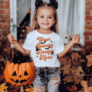 May include: A white t-shirt with a Halloween-themed design that says "Your Spooky Design Here" with bats, a witch's hat, and a spider. The shirt is being worn by a young child holding a pumpkin-shaped trick-or-treat bag.