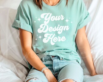 Puede incluir: Una camiseta azul claro con un gráfico blanco que dice "Your Design Here" en una fuente retro. La camiseta la lleva puesta una persona sentada en una cama blanca.