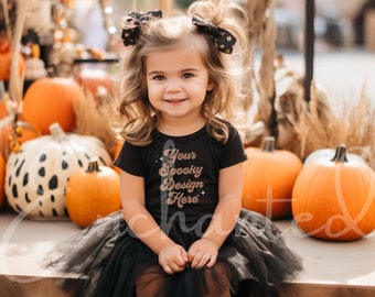 Può includere: Una bambina che indossa una maglietta nera con il testo "Your Spooky Design Here" in lettere dorate glitter. La bambina è seduta su una superficie di legno con una gonna di tulle nera e leggings marroni. La bambina è circondata da zucche.