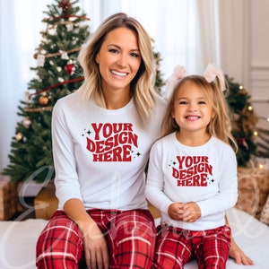 Op de afbeelding: Een moeder en dochter dragen bijpassende witte pyjama's met lange mouwen met rode tekst "YOUR DESIGN HERE". Ze dragen allebei rode en zwarte geruite pyjamabroeken. De afbeelding is gemaakt voor een kerstboom.