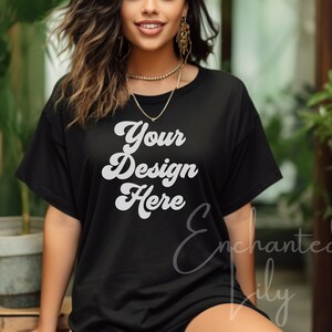 May include: A black oversized t-shirt with the text "Your Design Here" in white. The shirt is being worn by a person with long brown hair and is standing in front of a green plant.