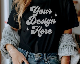 May include: A black t-shirt with the text "Your Design Here" in a white retro font. The shirt is being worn by a person with long blonde hair and is paired with blue jeans.