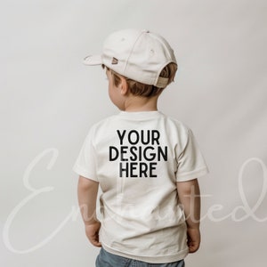 May include: A white t-shirt with short sleeves and the text "YOUR DESIGN HERE" printed on the back. The shirt is being worn by a child wearing a white baseball cap.