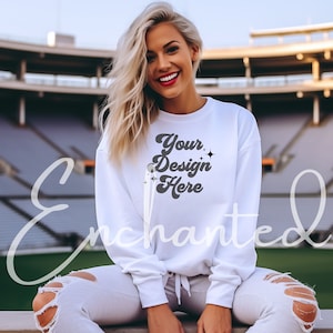 May include: White sweatshirt with the text "Your Design Here" in a retro font with sparkling stars. The sweatshirt is worn by a person sitting on a bleacher in a stadium.