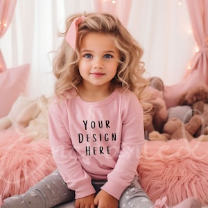 May include: A young girl wearing a pink sweatshirt with the text "YOUR DESIGN HERE" in black. She is sitting on a pink fluffy bed with stuffed animals in the background.