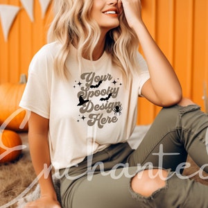 May include: A white t-shirt featuring a brown graphic design that reads "Your Spooky Design Here" with bats, a spider, and stars. The shirt is being worn by a person sitting on a bed of straw in front of a wall of orange pumpkins.