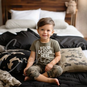 Pode incluir: Um menino pequeno vestindo uma camiseta verde com o texto "YOUR DESIGN HERE" sentado em uma cama com travesseiros de camuflagem.