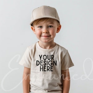 May include: A young child wearing a beige t-shirt with the text "YOUR DESIGN HERE" printed on the front. The child is also wearing a beige baseball cap.