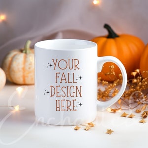 Peut inclure: Une tasse en céramique blanche avec un design textuel brun qui dit "Your Fall Design Here" avec de petites étoiles de chaque côté des mots. La tasse est posée sur une surface blanche avec des paillettes dorées et des citrouilles en arrière-plan.
